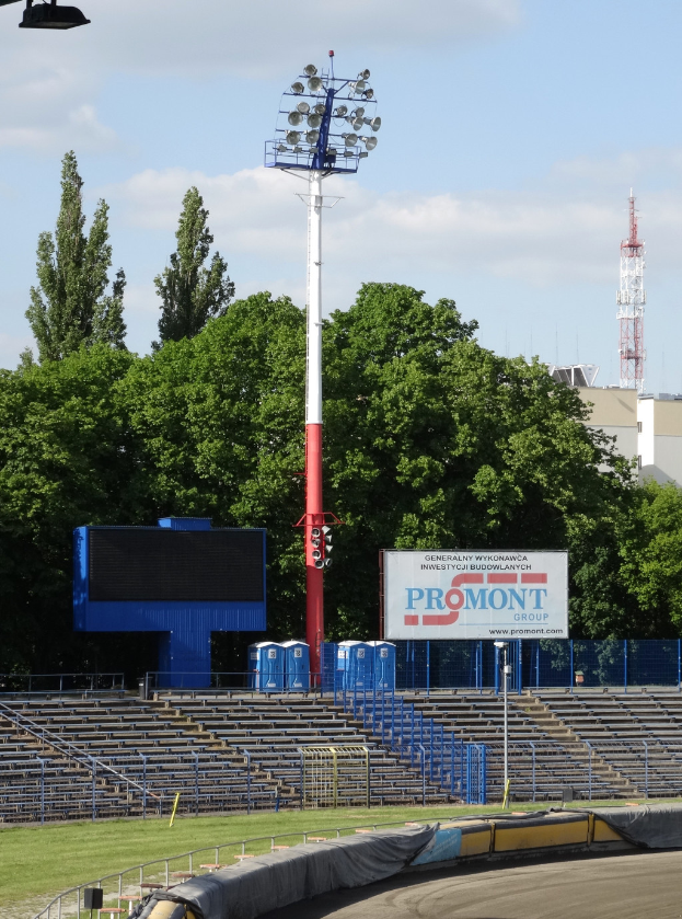 Eine von Scheinwerfern beleuchtete Rennbahn, umgeben von Gras, Geländern, Stufen, einem beleuchteten Pfahl, einem Schild, Bäumen, Gebäuden, Türmen und einem bewölkten Himmel.