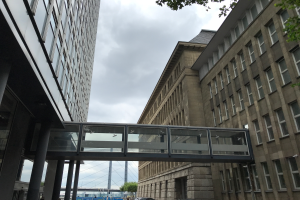 Ein Auto fährt eine Straße mit hohen Gebäuden entlang, mit einer Fußgängerbrücke in der Mitte, Menschen auf dem Bürgersteig und Bäumen, Pfählen und einem bewölkten Himmel im Hintergrund.