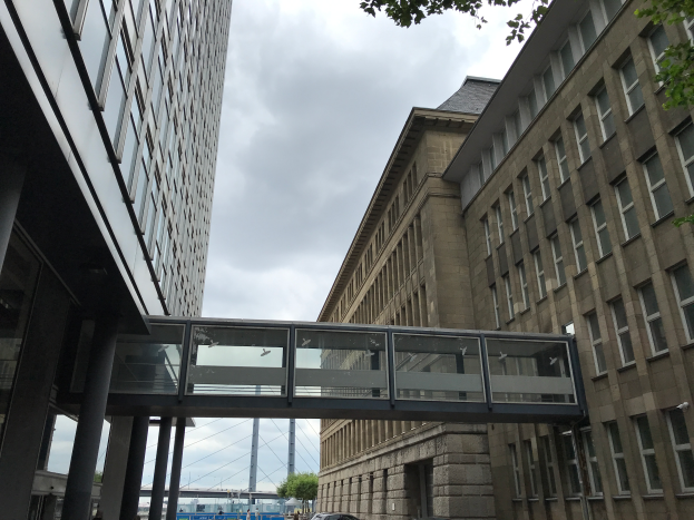 Ein Auto fährt eine Straße mit hohen Gebäuden entlang, mit einer Fußgängerbrücke in der Mitte, Menschen auf dem Bürgersteig und Bäumen, Pfählen und einem bewölkten Himmel im Hintergrund.