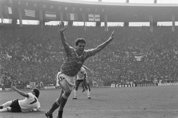 Schwarzes und weißes Foto eines Fussballspielers, der nach einem Tor in einem Stadion feiert, umgeben von Mannschaftskameraden und Mitarbeitern, mit Stadionelementen im Hintergrund.