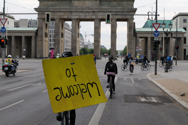 Eine Gruppe von Radfahrern fährt am Brandenburger Tor in Berlin, Deutschland vorbei, einer hält ein gelbes Schild; die Straße ist gesäumt von Laternenmasten und Gebäuden unter einem klaren Himmel.