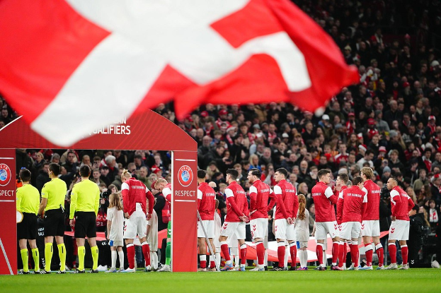 Eine Gruppe von Menschen auf einem Fuballfeld mit einer roten und weissen Flagge im Vordergrund, einem Bogen mit "Bayern Mnchen vs Bayern Mnchen Betting Tips & Preview" im Hintergrund und einer grossen Menge im Stadion.