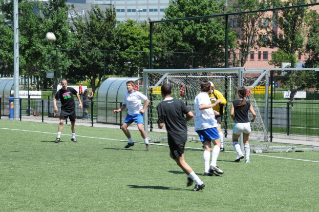 Menschen spielen Fußball auf einem Feld mit einem Zaun und einem Tor in der Mitte, einem Pfosten und einem Ball oben links und Bäumen und Gebäuden im Hintergrund.