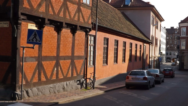 Fahrzeuge auf der Straße mit Gebäuden, Laternen, Verkehrszeichenmasten, Fenstern und Dächern im Hintergrund und ein Fahrrad auf dem Gehweg in der Nähe eines Pfahls.