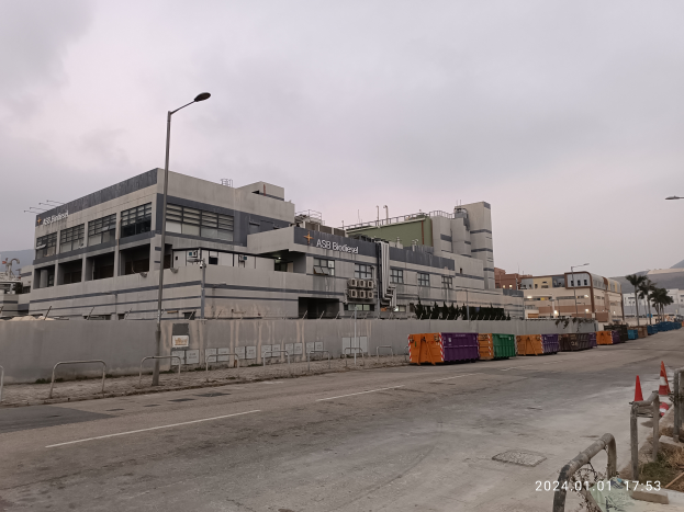 Ein großes Gebäude mit zahlreichen Fenstern, umgeben von Straßenmasten, Straßenlaternen, Absperrpollern, Verkehrskegeln, Containern, Bäumen, Hügeln und einem bewölkten Himmel, identifiziert als die neue Asb Bridgefeed-Einrichtung.