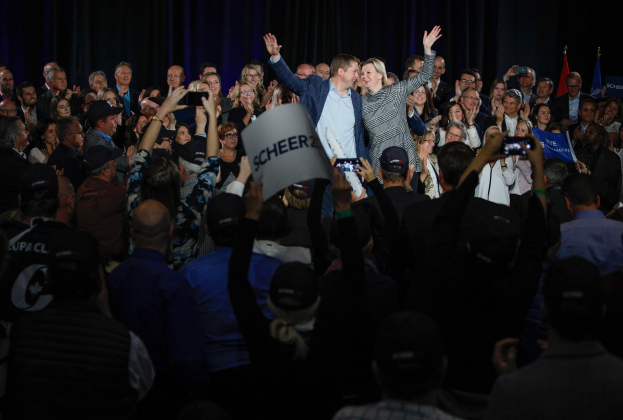 Ein Mann und eine Frau stehen vor einem Publikum, einige halten Schilder und Mobiltelefone, sowie Fahnen und einen Vorhang im Hintergrund.