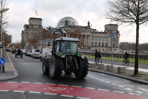 Ein grüner Traktor fährt am Reichstaggebäude in Berlin, Deutschland, vorbei, mit Passanten und Radfahrern auf dem Gehweg, Bäumen entlang der Straße und Flaggen am Gebäude.