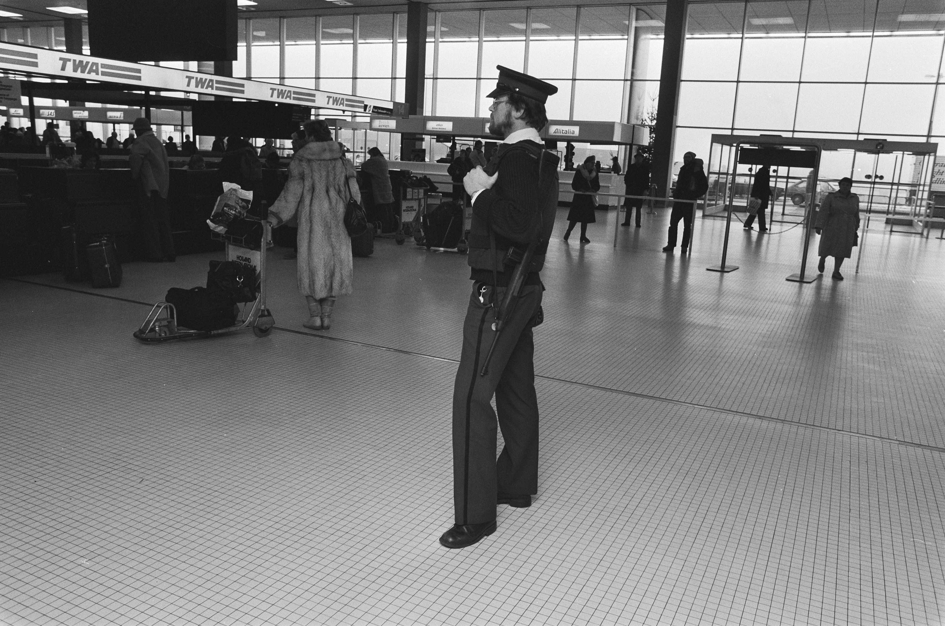 Polizeibeamter in Uniform mit einer Waffe in einem Flughafen-Terminal umgeben von Menschen und Gepäck.