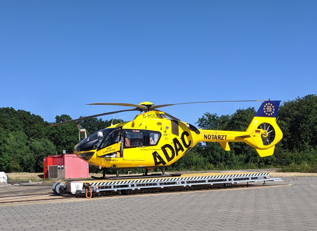 Ein gelber Hubschrauber sitzt auf dem Rollfeld eines Flughafens umgeben von Bäumen und einem klaren blauen Himmel, mit verschiedenen Gegenständen auf dem Rollfeld.