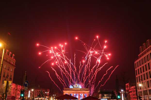 Eine Straßenansicht in Berlin an Silvester, mit Gebäuden, Bäumen, Laternenmasten, Ampeln, Schildern, Zelten und Menschen, und einem Feuerwerk am Himmel.