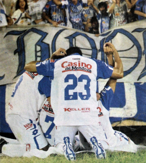 Eine Gruppe von Fußballspielern feiert ein Tor auf dem Feld mit einer jubelnden Menge im Hintergrund und einem teilweise sichtbaren Banner.