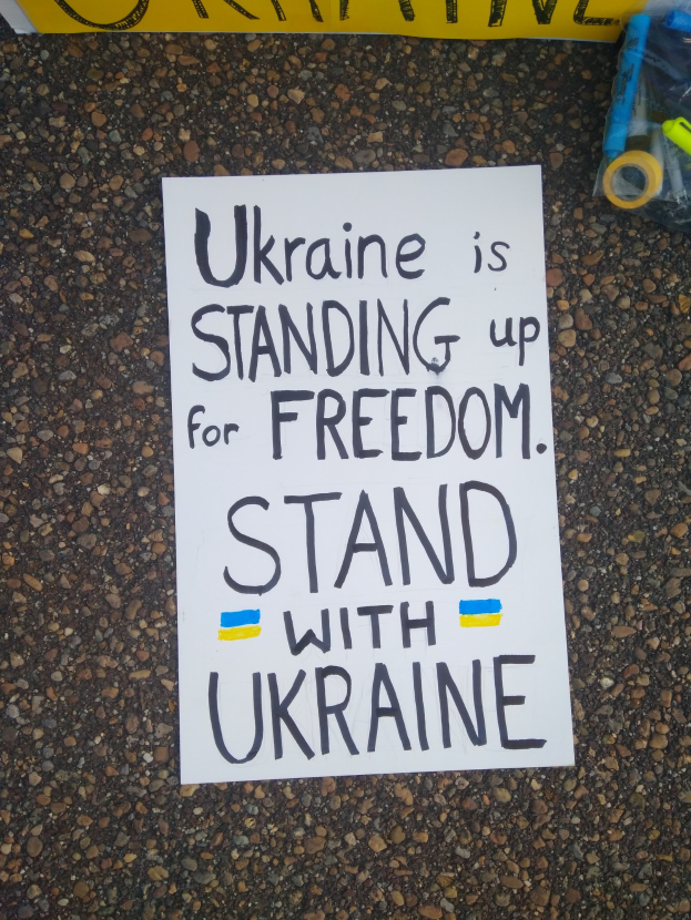 Ein weißer Zettel mit den Worten "Ukraine steht für Freiheit auf, stellt Euch mit Ukraine" liegt vor einer gelben Fahne auf dem Boden. Rechts daneben befindet sich eine Plastikabdeckung mit verschiedenen Gegenständen.