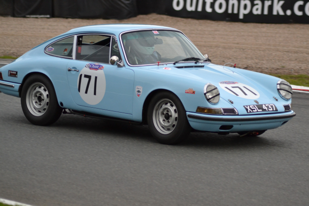 Blauer Porsche 911 GTS auf einer Rennstrecke mit einem Helmträger am Steuer, umgeben von Gras und einer schwarzen Fahne mit Text im Hintergrund.