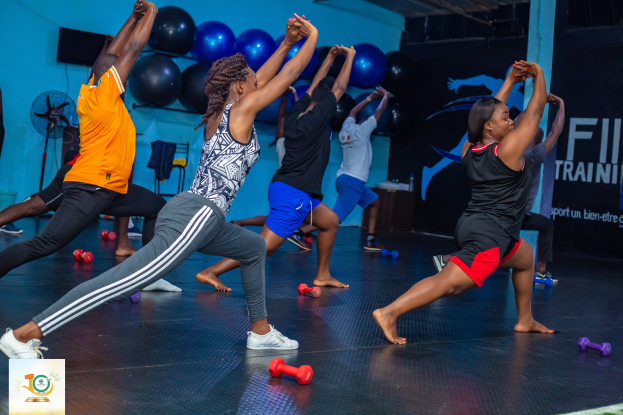 Eine Gruppe von Menschen, die Yoga in einem Fitnessstudio mit auf dem Boden verstreuten Hanteln machen, ein Banner im Hintergrund mit der Aufschrift "Fitness-Training in Ghana" und verschiedene Gegenstände wie Luftballons, ein Ventilator und ein Stuhl.