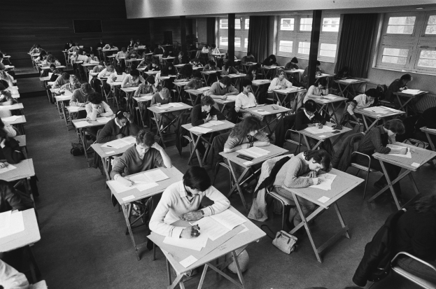 Ein Schwarz-Weiß-Foto einer Klasse mit Schülern an Tischen, die auf Papieren schreiben, mit Taschen auf dem Boden, Fenstern mit Vorhängen im Hintergrund und Lampen an der Decke.