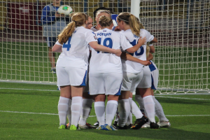 Eine Gruppe von Fussballspielerinnen in Uniformen steht auf dem Feld mit einem Netz und einer Person, die einen Ball hält, im Hintergrund.