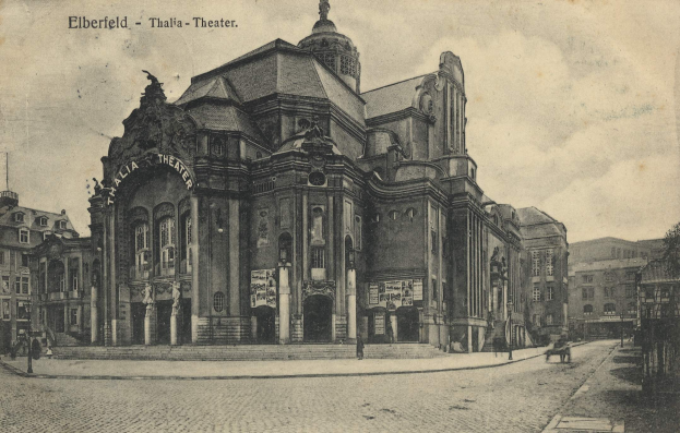 Ein altes Schwarz-Weiß-Foto des Elberfelder Theaters in Thalia, Deutschland, zeigt ein großes Gebäude mit Fenstern, Säulen, Bögen und Statuen, Menschen, die herumlaufen, eine Kutsche auf der Straße, Bäume im Hintergrund und einen bewölkten Himmel.
