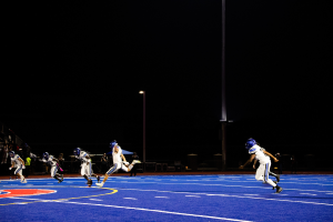 Eine Gruppe von Menschen in Sportkleidung und Helmen, die nachts Fußball spielen, mit Zuschauern im Hintergrund und Text unten auf dem Bild.