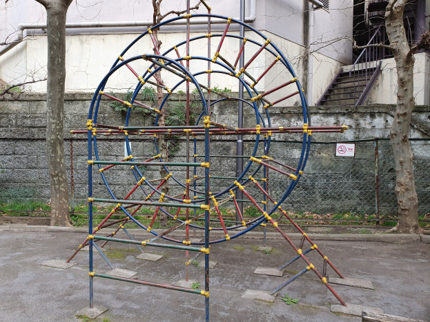Spielplatz mit großem Metallklettergerüst, umgeben von Bäumen und einer Mauer mit Maschendrahtzaun, mit Treppen mit Geländern, die zu einem Gebäude mit Fenstern und Rohren führen, ein Schild an einem Pfahl und grasbewachsenem Boden.