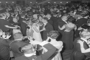 Ein Schwarz-Weiß-Foto einer Gruppe von Menschen auf einer Tanzfläche, einige halten Kameras, mit Treppen und Lichtern im Hintergrund.