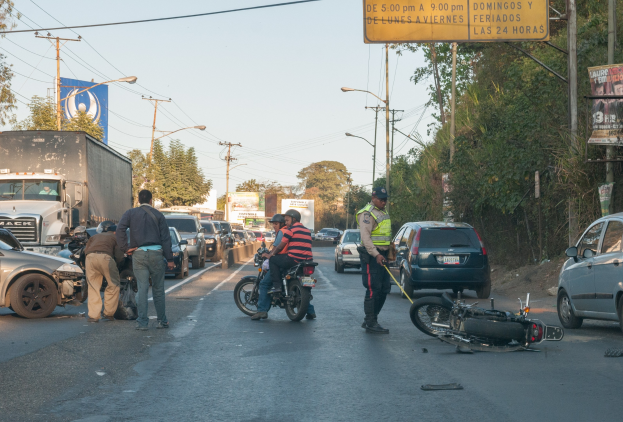 Gruppe von Menschen um ein verunglücktes Motorrad auf dem Seitenstreifen mit mehreren Fahrzeugen, darunter ein Lastwagen, im Hintergrund und Bäumen, Masten, Lichter, Schilder und Himmel.