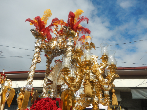 Ein großer goldener und roter Festwagen, geschmückt mit Blumen und anderen Dekorationen, in einem Karnevalsumzug, mit einem Gebäude, Strommasten mit Drähten und einem bewölkten Himmel im Hintergrund.