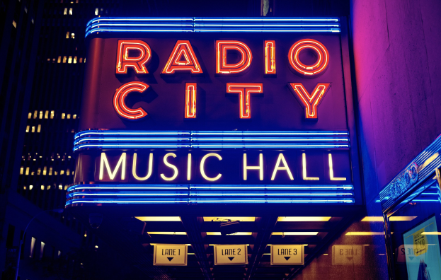 Außensicht des Radio City Music Hall in New York City, hell erleuchtet bei Nacht, mit hohen Gebäuden und einem "Radio City"-Schild auf der rechten Seite.