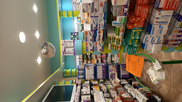 Eine Apotheken-Theke mit verschiedenen Medikamenten in Boxen und Flaschen auf Gestellen, mit einem Spiegel an der Wand, Deckenleuchten und einem Glastisch mit Papieren und Gegenständen darauf, der Boden ist unten sichtbar.