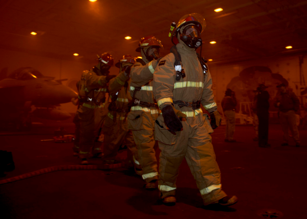 Eine Gruppe von Feuerwehrleuten in Schutzausrüstung durchquert eine Halle mit einem Flugzeug und verstreuten Gegenständen auf dem Boden.