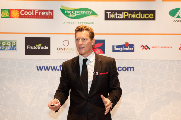 Ein Mann in einem schwarzen Blazer steht mit einem Gegenstand in der Hand und spricht, mit einem weißen Banner und einer weißen Tafel mit Organizational-Logos im Hintergrund.