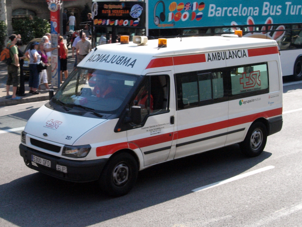 Rettungswagen fährt neben einem Bus die Straße entlang, mit Menschen auf dem Gehweg links neben dem Bus und Gebäuden, Bäumen und einem klaren blauen Himmel im Hintergrund.