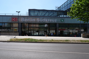 Ein großes Berliner Hauptbahnhof-Gebäude mit Glaswänden und Text, umgeben von Straßenlaternen, Autos, Menschen auf dem Gehweg, Fahrrädern, Bäumen und einem klaren blauen Himmel.