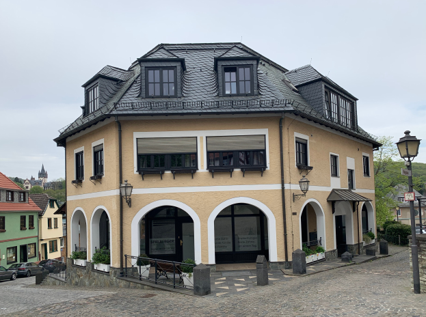 Ein großes Gebäude mit zahlreichen Fenstern in Heidelberg, Deutschland, umgeben von Straßenmasten, Fahrzeugen, Absperrpollern, Topfpflanzen, Schildern, Bäumen und einem bewölkten Himmel.