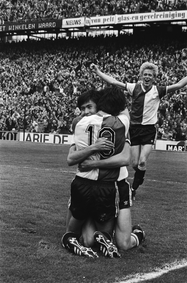 Drei Fussballspieler feiern ein Tor auf dem Boden, mit jubelnder Menge und schwenkenden Fahnen im Hintergrund, dargestellt in Schwarz-Weiss.