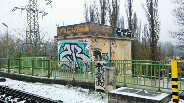 Eisenbahnschiene mit Graffiti an der Seite, umgeben von Zäunen, Pfosten, Lichtern und Bäumen, mit Türmen und Drähten im Hintergrund unter einem klaren Himmel und Schnee am Boden.
