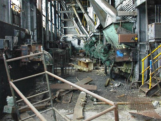Ein altes, ungenutztes Fabrikinnere mit Maschinen, Schutt auf dem Boden und Fenstern, die Geländer auf beiden Seiten aufweisen.