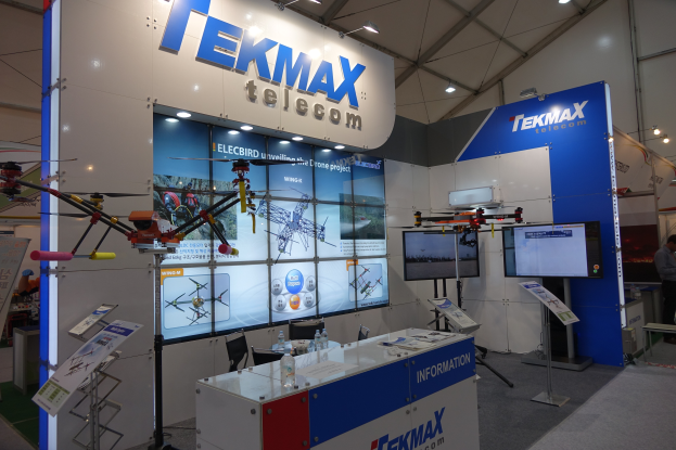 Eine Messestände für Tekmax Telecom mit einer großen Wandanzeige, Tisch mit Flaschen und Gegenständen, zwei Fernsehern, Texttafeln, Deckenleuchten und einigen stehenden und sitzenden Personen.