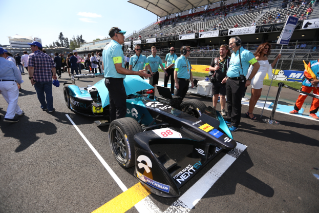 Eine Gruppe von Menschen feiert um ein Formel-E-Rennfahrzeug auf einer Strecke, mit einem Stadion voller Zuschauer im Hintergrund, unter einem klaren blauen Himmel.