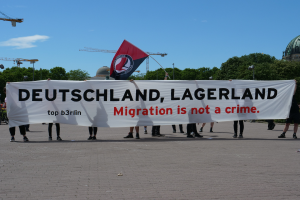 Eine Gruppe von Menschen, die eine Fahne mit der Aufschrift "Deutschland, Lagerland Migration ist kein Verbrechen" und eine Flagge halten, mit Straßenlaternen, Bäumen, Kränen, einem Gebäude und einem bewölkten Himmel im Hintergrund.
