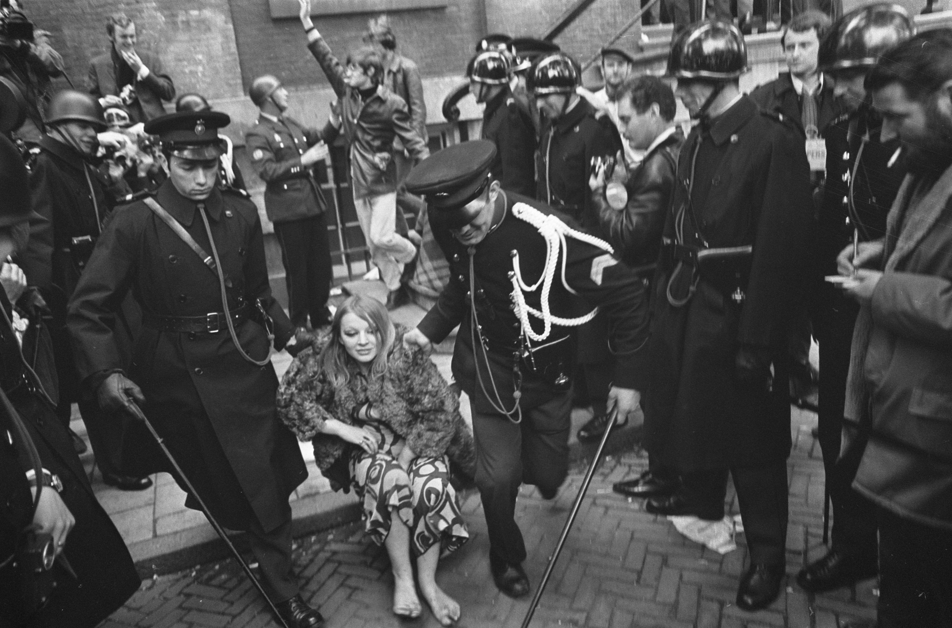 Schwarz-weißes Bild einer Frau, die von der Polizei an einer Menge mit Helmen vorbei geführt wird, die Stöcke halten, mit einem Gebäude mit Treppe und Geländer im Hintergrund.