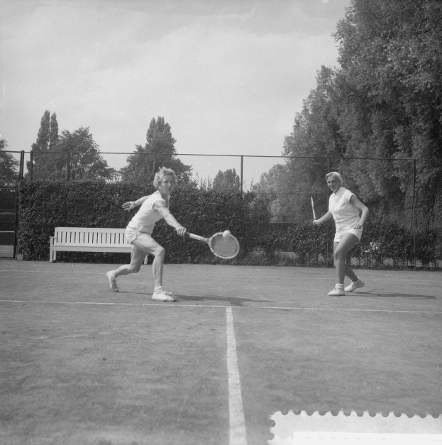 Zwei Frauen spielen Tennis auf einem Platz, jede hält einen Schläger in der Hand, mit einer Bank, einem Zaun, Pflanzen, Bäumen und einem klaren blauen Himmel im Hintergrund.