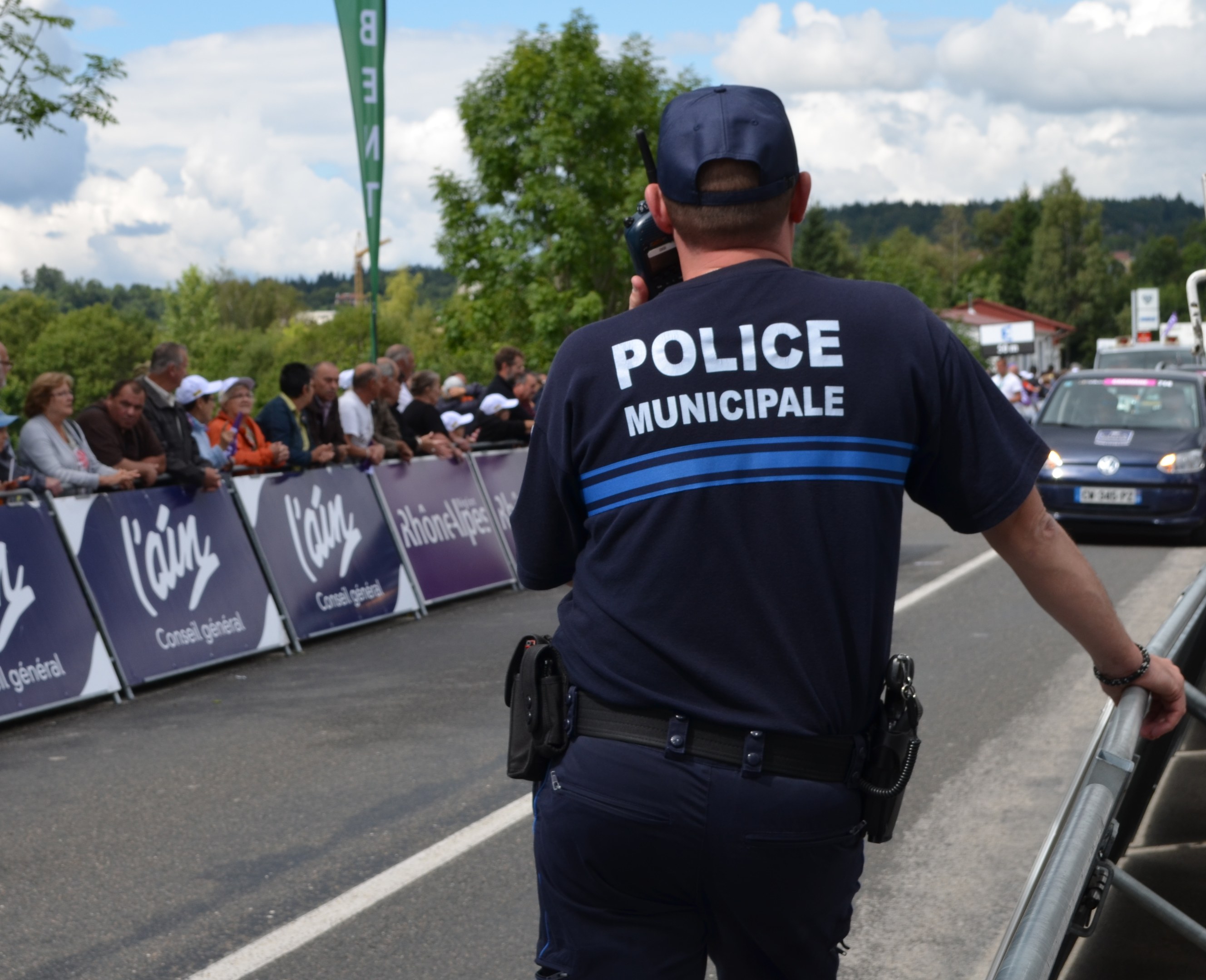 Polizeibeamter in Uniform mit Mütze und Funkgerät vor einer Menge mit Schildern, Fahrzeugen, Bäumen, Gebäuden und bewölktem Himmel im Hintergrund.