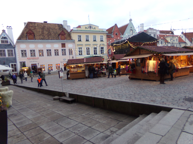 Ein lebendiger Weihnachtsmarkt in Tallinn, Estland, mit Menschen um dekorierte Stände, festliche Lichter und Gebäude mit Fenstern im Hintergrund bei einem bewölkten Himmel, mit Stufen, die zum Markt führen, und Töpfen mit Pflanzen, die herumstehen.