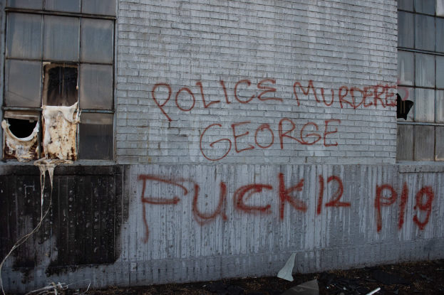 Graffiti an einer Gebäudewand mit der Aufschrift "Polizeimord George Fuck 12 P19" und eingeworfenen Fenstern und verstreuten Gegenständen davor.