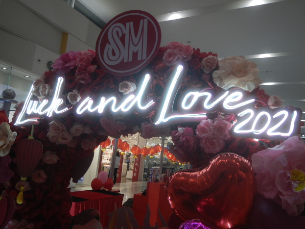 Ein Geschäft mit einem Schild, auf dem "Glück und Liebe 2021" steht, umgeben von Blumen, Ballons und anderen dekorativen Gegenständen, mit anderen Geschäften, Lichtern und einer Wand im Hintergrund, die eine festliche Atmosphäre für die SM-Valentinstag-Feier schaffen.