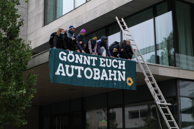 Eine Gruppe von Menschen auf dem Dach eines Gebäudes, die eine Fahne halten, mit einem Baum links und Glasfenstern im Hintergrund, wahrscheinlich protestierend für die deutsche Regierung.