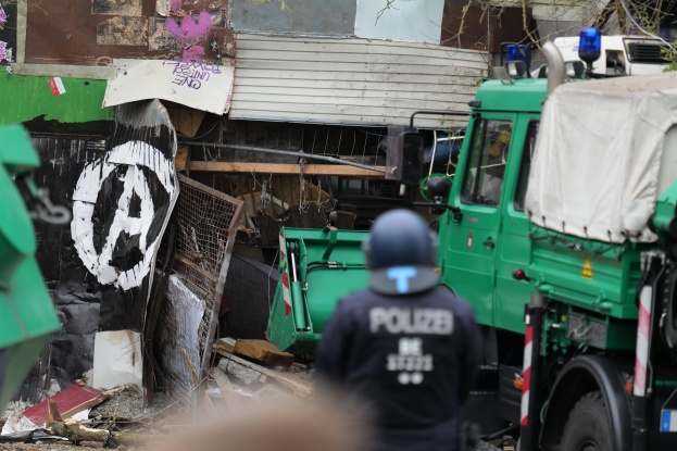 Ein Polizist steht vor einem Graffiti-bedeckten Gebäude, umgeben von Fahrzeugen, Bäumen und anderen Gegenständen, mit einem menschlichen Gesicht am unteren Bildrand.