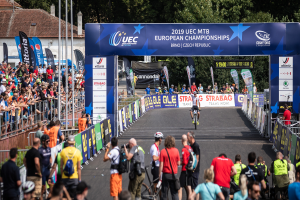 Eine Gruppe von Radfahrern fährt eine Straße entlang, die von Zuschauern gesäumt ist und von einem Bogen gekrönt wird, auf dem "2019 UEC MTB Europameisterschaften" steht, mit Bäumen, Gebäuden und einem klaren blauen Himmel im Hintergrund.