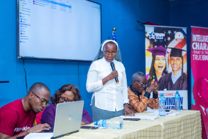 Frau spricht in ein Mikrofon auf einer Besprechung mit Menschen, die um einen Tisch mit Laptops und Papieren sitzen, mit einem Banner und Flaggen im Hintergrund.