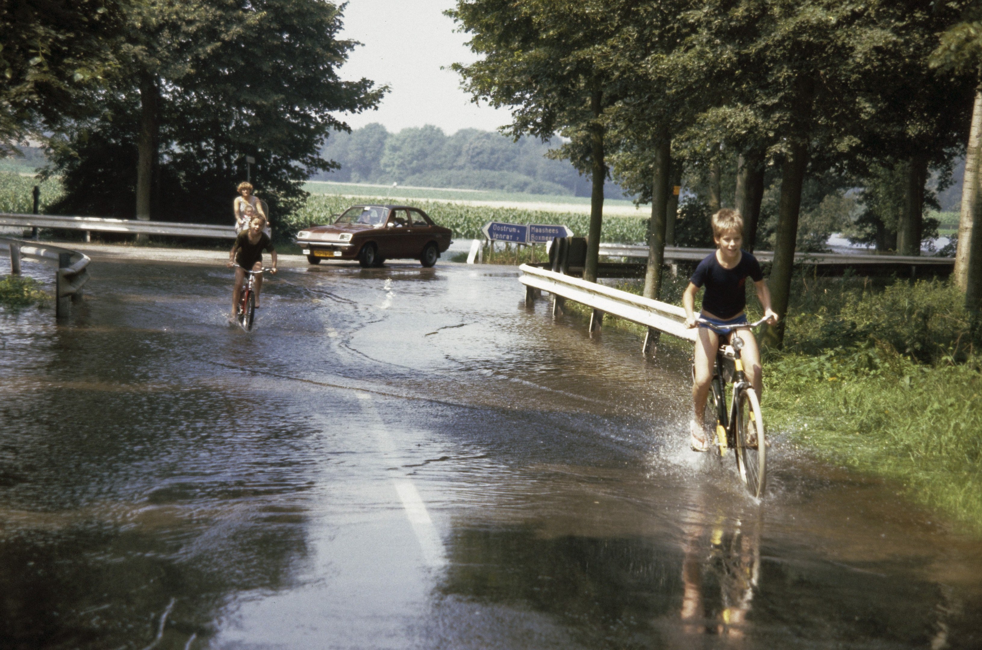 Eine Gruppe von Menschen fährt mit Fahrrädern eine überflutete Straße hinunter, mit einem daneben fahrenden Auto, gesäumt von einem Geländer und umgeben von Bäumen und Pflanzen, unter einem klaren blauen Himmel.